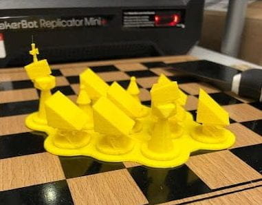 A set of yellow chess pieces featuring large wedges of cheese on top sitting on a wooden chessboard in front of the 3D printer.