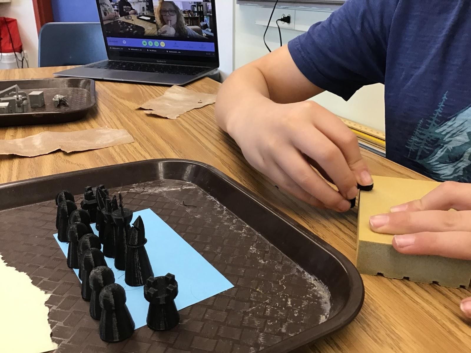 A student's hands sanding a black chess piece next to a tray with the other chess pieces from the set. 