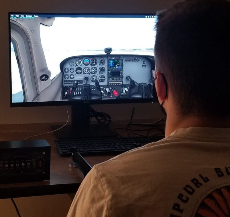 A student sitting at a computer with a flight simulator screen. Looking over his shoulder the screen shows the cockpit of a small plane.
